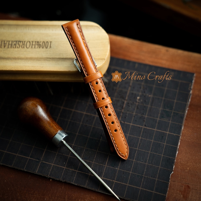 May include: A brown leather watch strap with stitching detail. The strap is lying on a brown grid surface with a wooden block and a leather awl.