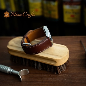 May include: A brown leather watch strap with white stitching is shown on a wooden brush. The strap is on a silver watch case. The brand name "Mino Crafts" is visible in the top left corner of the image.