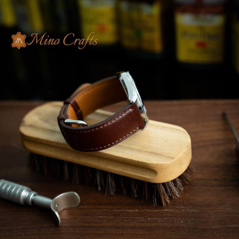 May include: A brown leather watch strap with white stitching is shown on a wooden brush. The strap is on a silver watch case. The brand name "Mino Crafts" is visible in the top left corner of the image.