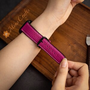 May include: A pink leather watch strap with white stitching and a black buckle. The strap is shown on a person's wrist.