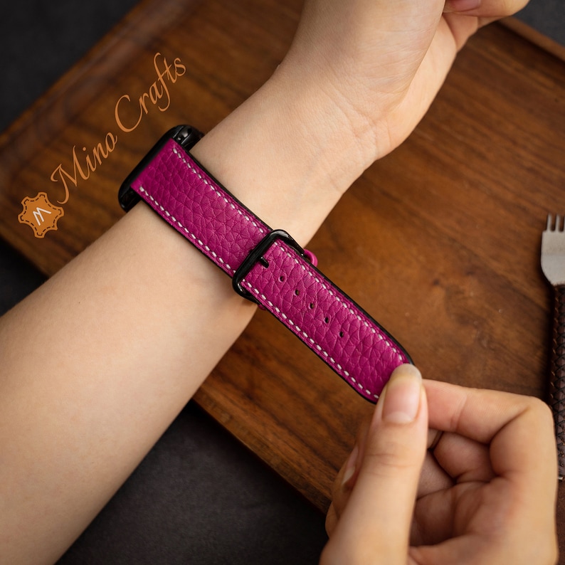 May include: A pink leather watch strap with white stitching and a black buckle. The strap is shown on a person's wrist.