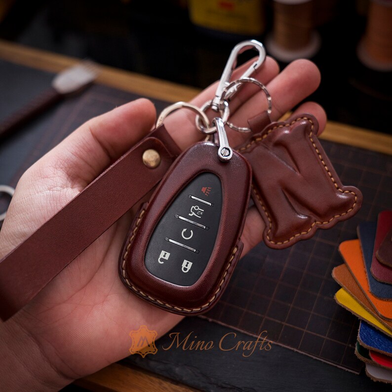 Op de afbeelding: Een bruine leren sleutelhanger met een zilveren sleutelring en een bruine leren hanger met een initial. De sleutelhanger heeft een gestikt ontwerp en is bevestigd aan een bruine leren band.