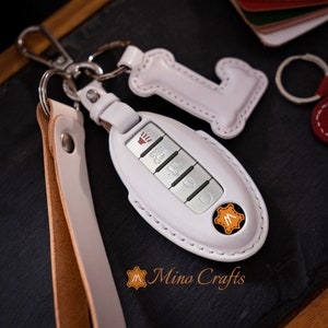 May include: White leather key fob with a silver key and a small white leather charm shaped like the letter L. The key fob has a brown leather strap attached to it. The key fob is labeled "Mino Crafts".
