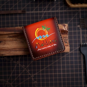 May include: A brown leather wallet featuring the word "Maldives" and a tropical design. The wallet has a stitched border and the text "...the sunny side of life".