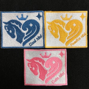 以下が含まれることがあります： 白い背景に、王冠と星をつけた馬が描かれたハート型のデザインの刺繍パッチが3つあります。パッチは青、黄、ピンクで、それぞれ「Cool Idol」、「Classic Idol」、「Cute Idol」と書いてあります。
