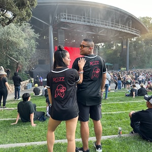 May include: Black baseball-style jerseys with white trim and the word "REZZ" in white block letters. A red graphic of a person wearing sunglasses and a hat is on the back. The jerseys are worn by two people at an outdoor concert.