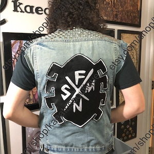 以下が含まれることがあります： 白いスタイリッシュなロゴに「SF」の文字と打ち消された「Z」が描かれた黒いパッチが付いた青いデニムベスト