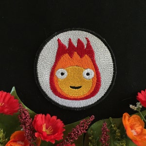 May include: Embroidered patch featuring a cartoon fire spirit with a smiling face, red and orange flames, and white eyes. The patch is circular and has a white background with a silver border.