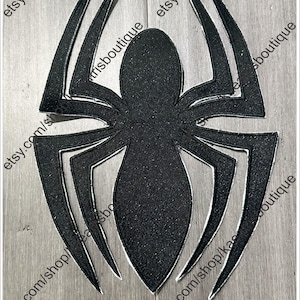 Könnte beinhalten: Ein schwarzes Spinnenemblem mit glitzernder Textur und weißer Umrandung. Die Spinne hat acht spitze Beine und einen großen ovalen Körper. Der Hintergrund ist ein hellgraues Holzmaserung.