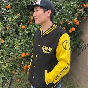 Puede incluir: Chaqueta universitaria negra y amarilla con el texto "CL4P-TP" y "Claptrap". La chaqueta tiene mangas amarillas y un cuerpo negro con rayas amarillas. Se lleva una gorra de béisbol negra.