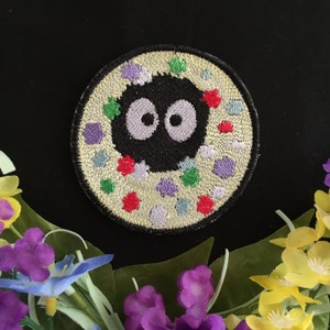 May include: A black and white embroidered patch of a round, white, and colorful creature with two large eyes. The creature is surrounded by a circle of colorful dots.