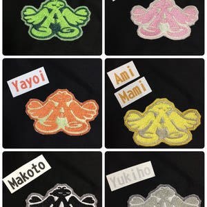 以下が含まれることがあります： 腕を広げた漫画のキャラクターが描かれた、アイロン接着式の刺繍パッチが5つ。パッチは緑、ピンク、オレンジ、黄色、グレーなど、さまざまな色があります。パッチには、上に白いラベルで名前が書かれています。ミキ、ロリ、ヤヨイ、アミマミ、ユキホです。