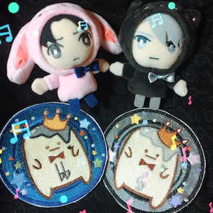 May include: Two plush toys, one pink bunny and one black cat, with embroidered faces.  The bunny is wearing a pink hoodie with a bow tie. The cat is wearing a black hoodie with a bow tie.  Two embroidered patches with a white character wearing a crown and bow tie on a blue and gray background.
