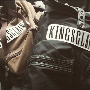 May include: Two black and brown duffel bags with the word "KINGSLAYER" printed in white on one bag and "SLAYER" printed in white on the other bag.