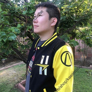 May include: Black and yellow varsity jacket with a button-up front. The jacket has yellow sleeves and a black body with white and yellow stripes on the collar and cuffs. The left sleeve has a circular logo, and the front has a design with the number 11.