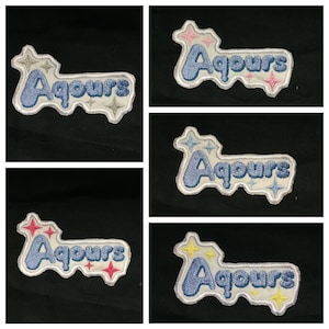 Puede incluir: Cuatro parches bordados con el texto "Aqours" en azul con contornos blancos. Cada parche tiene un diseño de estrella de color diferente. Las estrellas son rosas, blancas, amarillas y rojas.