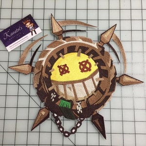 May include: Embroidered patch featuring a smiling, circular character with a yellow face, brown accents, and a chain. The character has cross-shaped eyes and a wide grin. The patch has several pointed extensions and a small green detail.