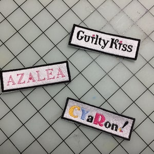 May include: Three embroidered patches with white backgrounds and black borders. The top patch says "Guilty Kiss" in black lettering. The middle patch says "AZALEA" in pink lettering. The bottom patch says "CYaRoN!" in yellow, red, and blue lettering.