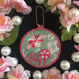 May include: A round, green embroidered patch with pink stitching. The patch features a pink and white candy cane, a spool of thread, a needle and thread, and a pink flower. The word "Ruby" is embroidered in pink on the patch.