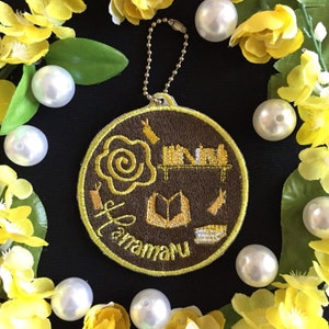 Könnte beinhalten: Ein brauner und gelber bestickter Schlüsselanhänger mit dem Wort "Haramaru" und Bildern von Büchern, einem Bücherregal und einer Rose.
