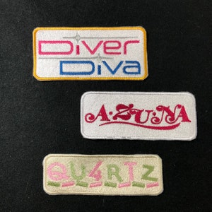 May include: Three embroidered patches with white backgrounds and colorful lettering. The top patch is yellow with the text "Diver Diva" in pink, blue, and white. The middle patch is white with the text "A:ZUNA" in red. The bottom patch is cream with the text "QUARTZ" in pink and green.
