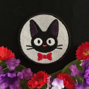 May include: A black embroidered patch of a cat wearing a red bow tie. The cat has large, white eyes and a small, pink nose. The patch is circular and has a white background.