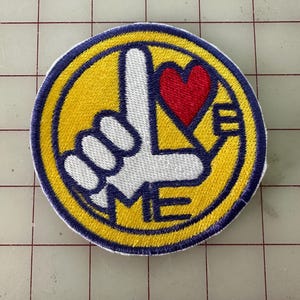 Puede incluir: Un parche circular bordado de color amarillo y azul con una mano blanca haciendo el gesto de 'Te amo'. La mano sostiene un corazón rojo. El texto 'ME' está bordado en azul debajo de la mano.