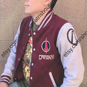 May include: Maroon and gray letterman jacket with white accents. The jacket features a Crimson Lance logo and text on the front, and a black symbol on the sleeve. The jacket has a button-up closure and striped cuffs.