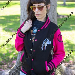 Peut inclure: Veste universitaire noire et rose avec une fermeture boutonnée sur le devant, avec un motif sur la poitrine et la manche. La veste a des manches, des poches et des bordures roses, avec des rayures noires sur les poignets.