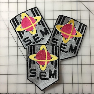 以下が含まれることがあります： 赤い星と黄色の星をスタイリッシュにデザインした、銀と黒の刺繍パッチが3つ。テキストは「S.E.M.」