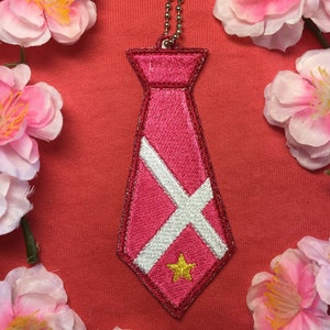 May include: A pink tie-shaped keychain with a white X and a gold star. The keychain is hanging from a silver chain.