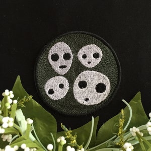 May include: A dark green embroidered patch featuring four white faces with black eyes. The faces are arranged in a circle.