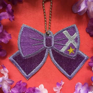 Puede incluir: Un parche bordado morado en forma de lazo con un diseño de cruz plateada y estrella amarilla. El parche tiene una cadena plateada adjunta para colgar. El fondo es de color coral, rodeado de flores moradas.