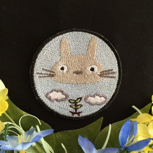 May include: Embroidered patch featuring a cartoon Totoro character with a light blue background and white clouds. The character is brown with a light brown nose and large eyes. A small green plant is in the center of the patch.