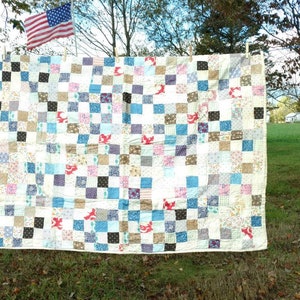 May include: A patchwork quilt with a checkered pattern in shades of white, blue, brown, and pink. The quilt is hanging on a clothesline outdoors.