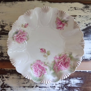 May include: A white porcelain serving dish with a scalloped edge and a floral design featuring pink roses. The dish has a raised rim and a smooth, glossy finish.