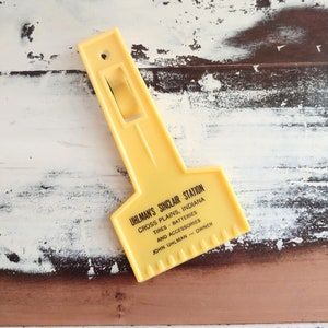 May include: A yellow plastic ice scraper. The scraper has a handle and a notched edge. The text "UHLMAN'S SINCLAIR STATION" is printed on the scraper, along with the location and owner's name.