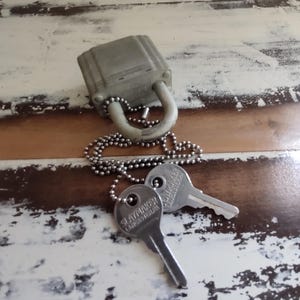 Puede incluir: Un candado gris desgastado con una cadena plateada y dos llaves. Las llaves están marcadas con "SLAYMAKER LANCASTER PA". El candado y las llaves descansan sobre una superficie de madera envejecida, blanca y marrón.