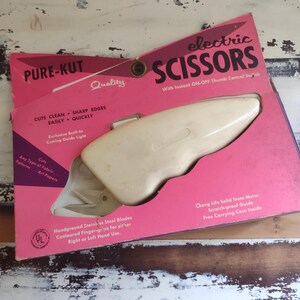 Könnte beinhalten: Vintage elektrische Schere in einer rosa Schachtel. Auf der Schachtel steht "PURE-KUT" und "electric SCISSORS". Die Schere ist cremefarben und hat einen Daumenschalter. Die Schachtel gibt an, dass die Schere jede Art von Stoff schneidet.