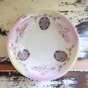 May include: A white ceramic bowl with a pink and white floral design. The bowl has a scalloped edge and a raised rim.