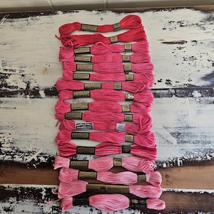 May include: A collection of fourteen skeins of J. & P. Coats six-strand embroidery floss arranged in a vertical column. The threads feature a gradient of pink and red shades, ranging from deep crimson at the top to light pastel pink at the bottom.