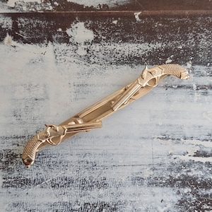 Puede incluir: Pinza de corbata dorada con un diseño detallado de dos pistolas antiguas. La pinza tiene una barra rectangular con los diseños de pistolas en cada extremo, mostrando texturas y formas intrincadas. El fondo es una superficie clara y desgastada.