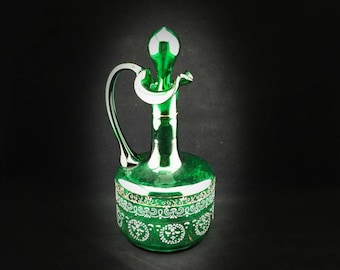 May include: A green glass decanter with a white and gold design. The decanter has a wide base and a narrow neck with a curved spout.
