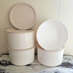 May include: A set of round storage containers with white bodies and beige lids. The containers feature a smooth finish, with one lid standing upright behind the stack and one container positioned to the right with its interior clearly visible.