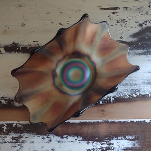 May include: A dark iridescent glass bowl with a wavy, scalloped edge. The bowl has a circular, multi-coloured design in the centre, with shades of green, blue, pink, and purple.