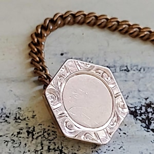 May include: Close-up of a vintage gold-toned locket with an octagonal shape and a decorative chain. The locket features an engraved design around a central, circular area. The chain is a twisted link style.
