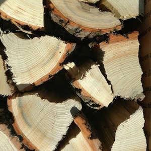 May include: Close-up of a pile of split firewood. The wood pieces have light brown and tan tones with visible growth rings. The edges of the wood have a darker brown bark.