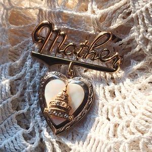 Op de afbeelding: Goudkleurige broche met het woord "Mother" in cursief boven een hartvormige hanger. De hanger heeft een witte parelmoeren kern en een goudkleurig gebouw met heldere strass-steentjes. De broche is geplaatst tegen een crèmekleurige, gehaakte achtergrond.