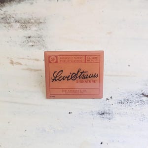 May include: A rectangular, salmon-colored Levi Strauss & Co. label with black text. The label reads "AUTHENTIC PATENT RIVETED CLOTHING" and "Levi Strauss SIGNATURE." The label also includes the text "U.S. PATENT MAY 20, 1873" and "San Francisco, Cal."