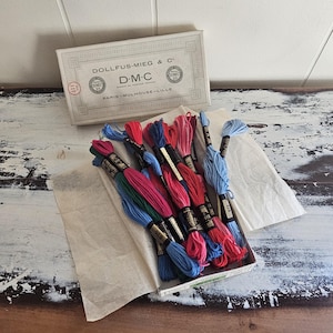 May include: A collection of multi-colored embroidery floss skeins in a cardboard box labeled DOLLFUS-MIEG & C D.M.C PARIS-MULHOUSE-LILLE. The threads are in shades of red, pink, blue, and green, each wrapped with a black paper band featuring gold text.
